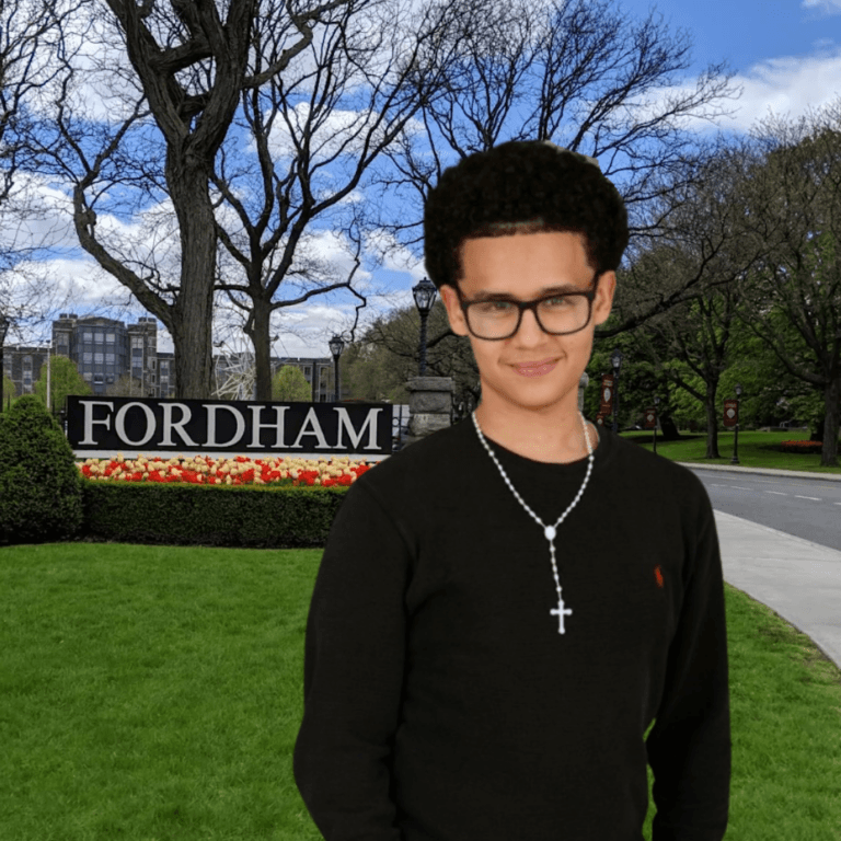 Young person with glasses and a cross necklace stands on a lawn in front of a Fordham sign and campus landscaping, near the Jeffrey Gural Student Commons.