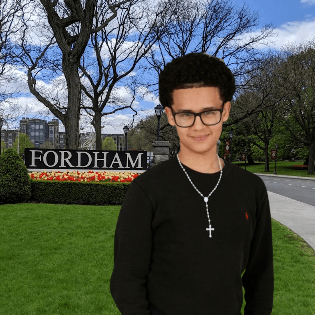 Young person with glasses and a cross necklace stands on a lawn in front of a Fordham sign and campus landscaping, near the Jeffrey Gural Student Commons.
