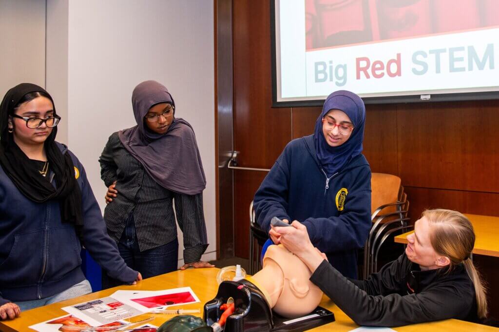 Four students participate in a medical simulation using a mannequin arm in a classroom for a STEM exposure program