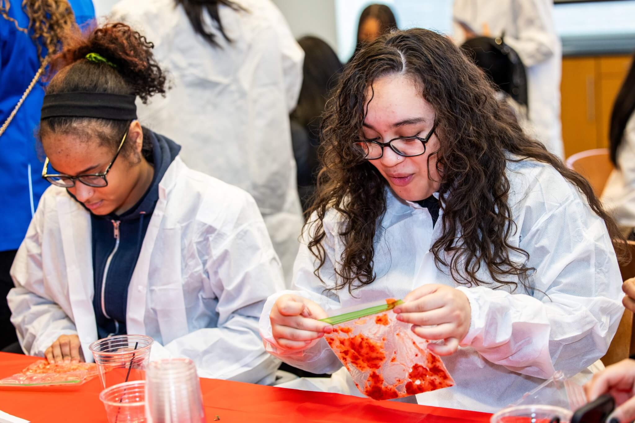 Two people wearing white lab coats work at a table with plastic measuring cups and a bag of crushed fruit