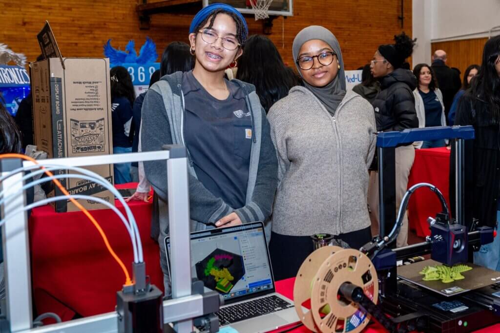 Two students stand behind a table with 3D printers, a laptop displaying a colorful model, and filament spools at a science fair STEM exposure event