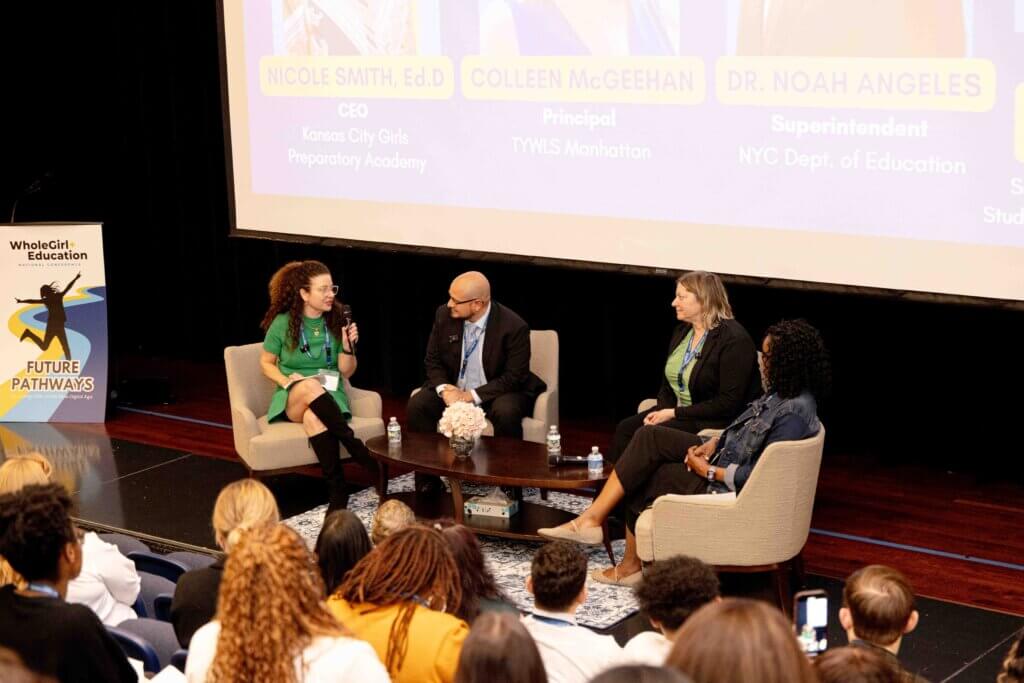 Four panelists sit and speak onstage at an education conference, with an audience watching and a presentation slide—featuring their names, titles, and highlights from the Annual Report 2025—displayed behind them.