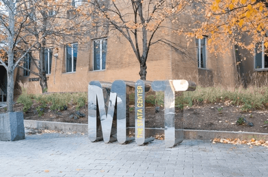 Large metallic MIT letters, with The Stata Center written vertically on the I, stand in front of a brick building and autumn trees near the Jeffrey Gural gateway.