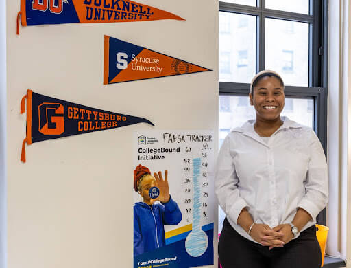 A woman stands smiling by a wall adorned with college pennants, a FAFSA tracker chart, and a Jeffrey Gural poster in a brightly lit room.