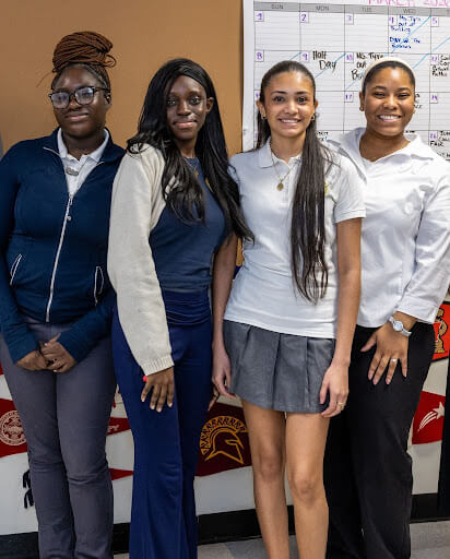Four students, including Jeffrey Gural, stand side by side indoors, smiling at the camera. A whiteboard calendar and posters are visible in the background.