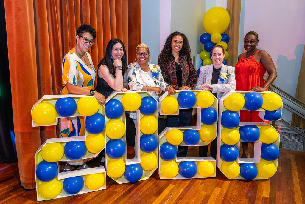 Six people stand behind large 2026 numbers filled with blue and yellow balloons; balloons and curtains decorate the background.