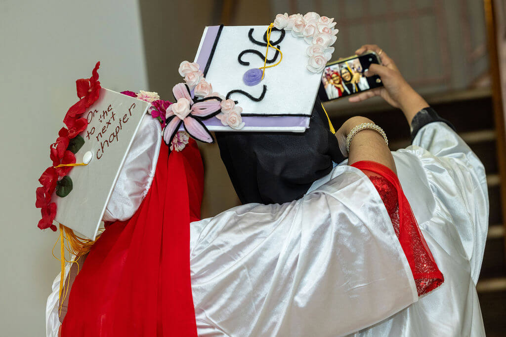 Two graduates in decorated caps and hijabs take a selfie together; one cap reads On to the next chapter, the other has 2026 and floral decorations.