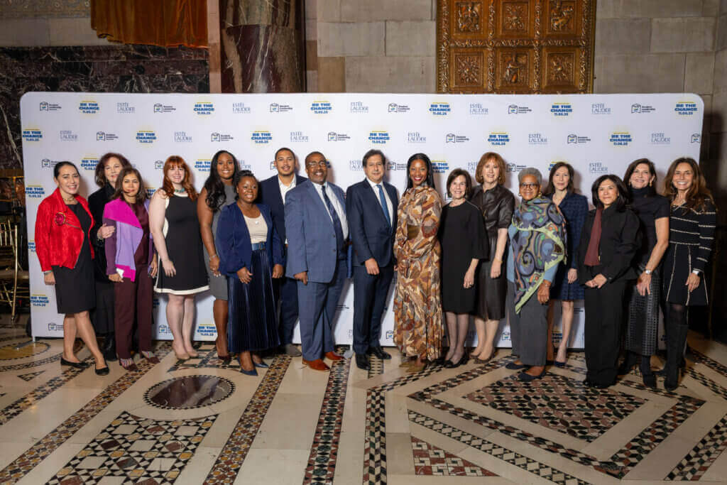 A group of people in formal attire pose for a photo on a patterned floor in front of a branded step-and-repeat banner at the Annual Report 2025 indoor event.