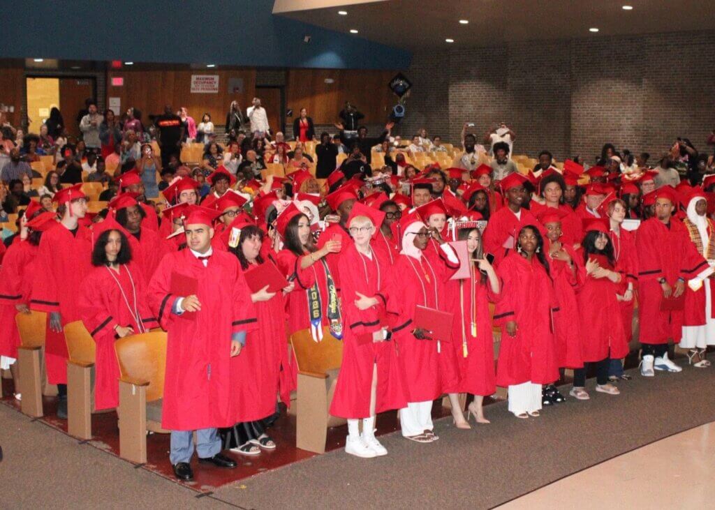 A large group of students in red graduation gowns and caps stand and pose for photos in an auditorium filled with people.