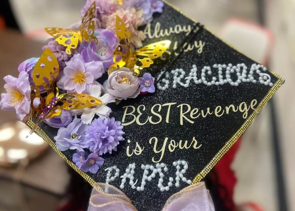 A graduation cap decorated with artificial purple flowers, gold butterflies, and text reading “BEST Revenge is Your PAPER” in gold and white letters.