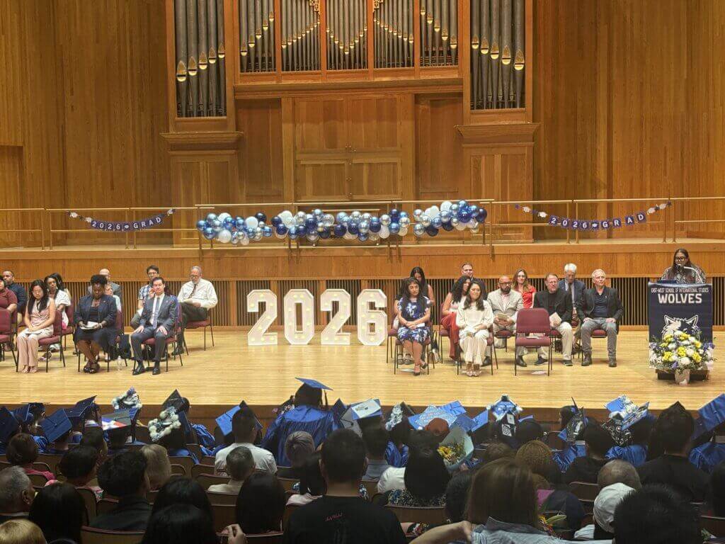 A graduation ceremony in an auditorium features students in caps and gowns, a stage with 2026 numbers, seated speakers, balloons, and an audience.