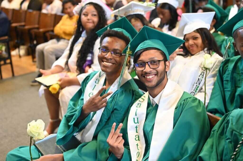 Two Class of 2026 graduates in green caps and gowns sit and smile at the camera, making peace signs.
