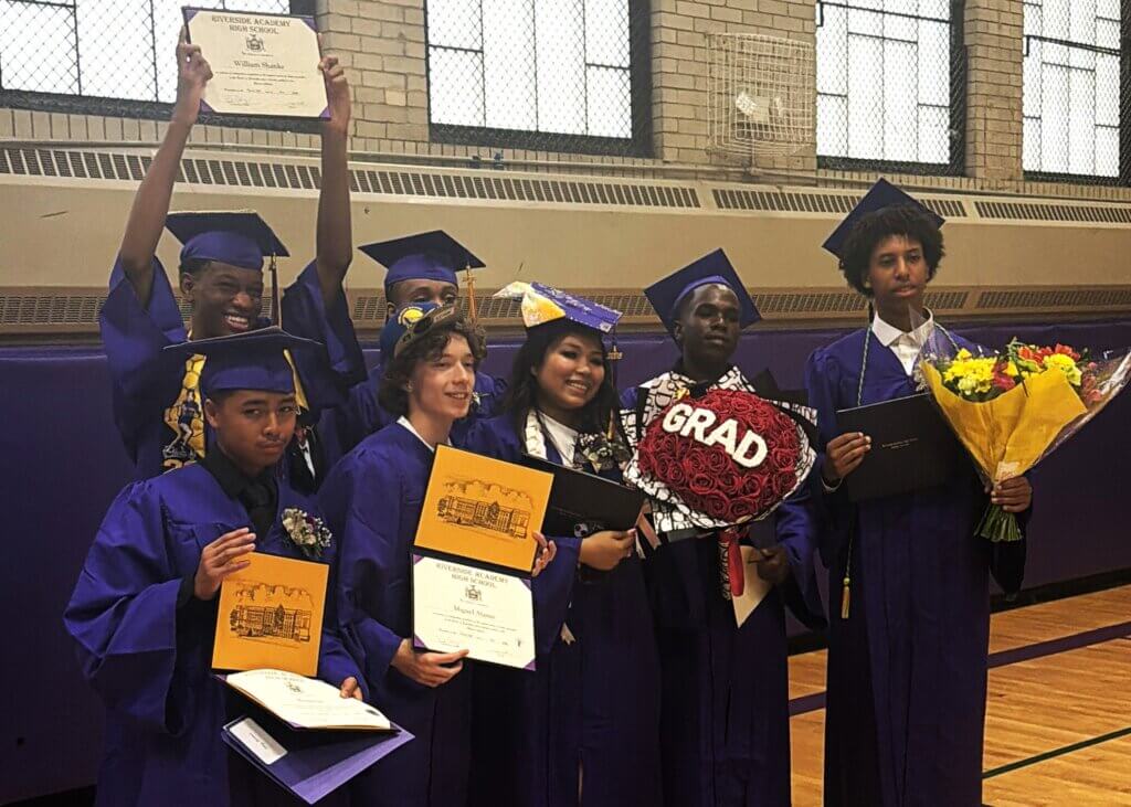 Seven graduates in purple gowns and caps pose indoors, holding diplomas, a bouquet, and a GRAD sign. Some are smiling and one raises a certificate above his head.