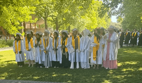 A group of graduates in white caps and gowns with gold stoles stand in a row outside on a sunny day, posing for a photo.