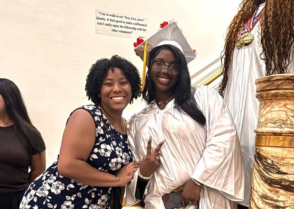 Two women pose and smile; one is wearing a white graduation cap and gown, holding up a peace sign, while the other wears a navy floral dress. A quote is visible on the wall behind them.