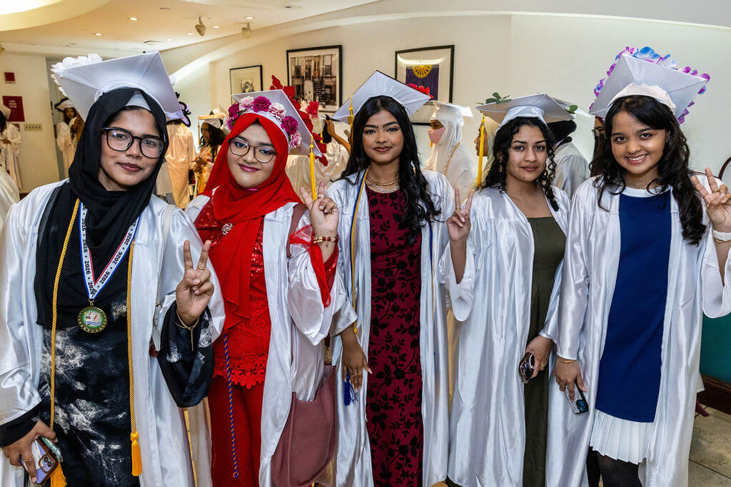 Five graduates in white gowns and caps stand indoors, smiling and making peace signs, with others in similar attire visible in the background.