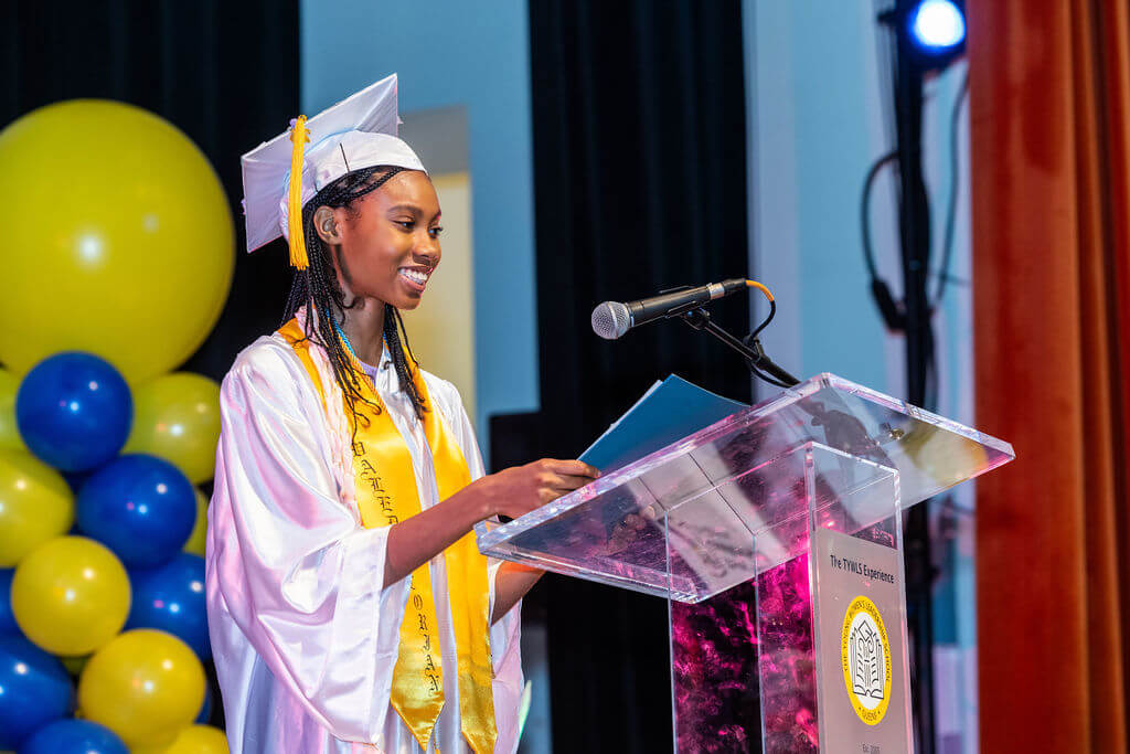 A young woman in a graduation cap and gown stands at a podium, smiling while delivering a speech at a ceremony with balloons in the background—her confidence and poise reminiscent of an Estée Lauder ambassador.