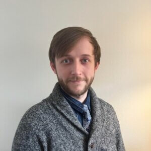 Devin Chowske, the teacher who led TYWLS Astoria's AI policy process, is standing in front of a plain wall. He is wearing a grey sweater and blue tie.