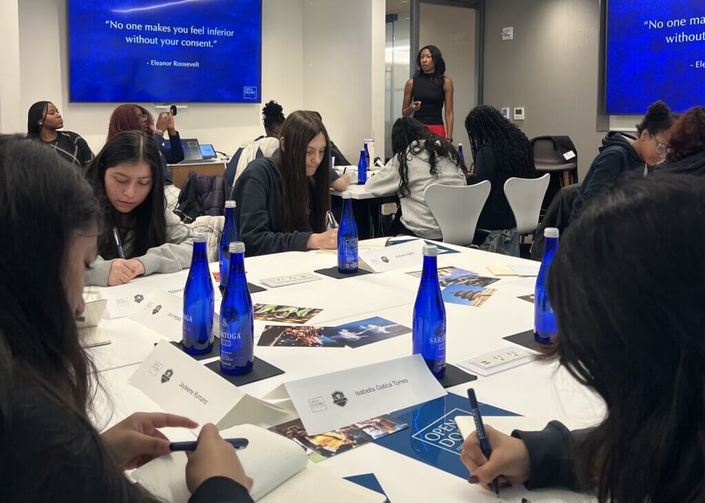 A group of students work at a table while a woman presents near a screen displaying an Eleanor Roosevelt quote in a modern classroom setting.