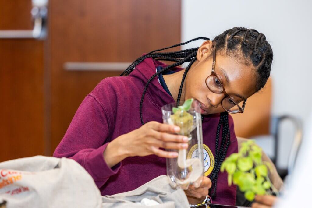 A person wearing glasses and a maroon hoodie closely examines a plant specimen inside a clear container in an indoor setting.