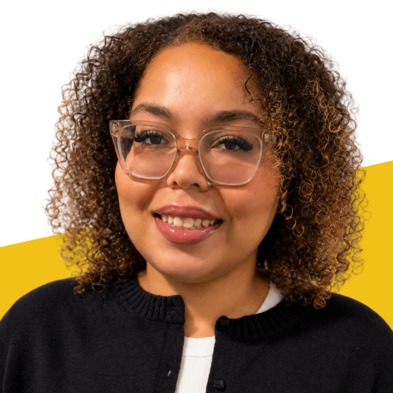 Nashalie Robledo, a woman with curly hair and glasses, smiles at the camera. She is wearing a black sweater over a white top, with a yellow and white background behind her.