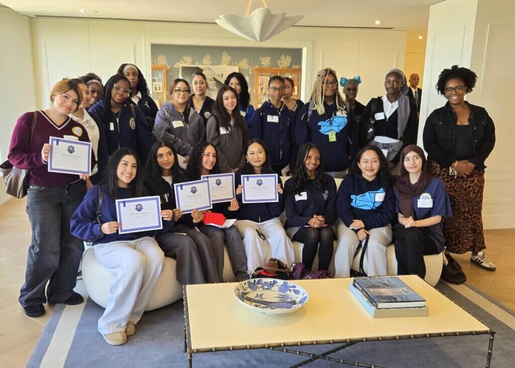 A group of women and girls pose indoors, some holding certificates, with a few seated in front and the rest standing behind them.