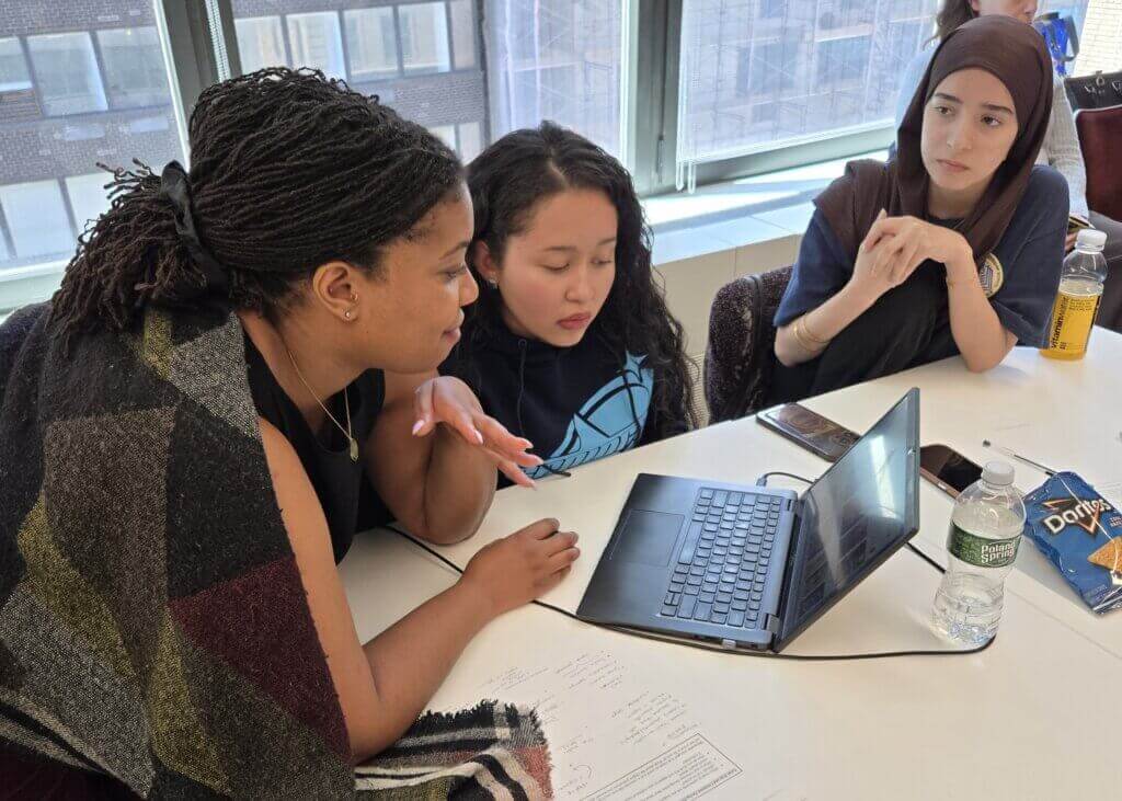 Three women sit at a table, focusing on a laptop. Papers, snacks, and drinks are spread out as they work together in a brightly lit room with large windows.