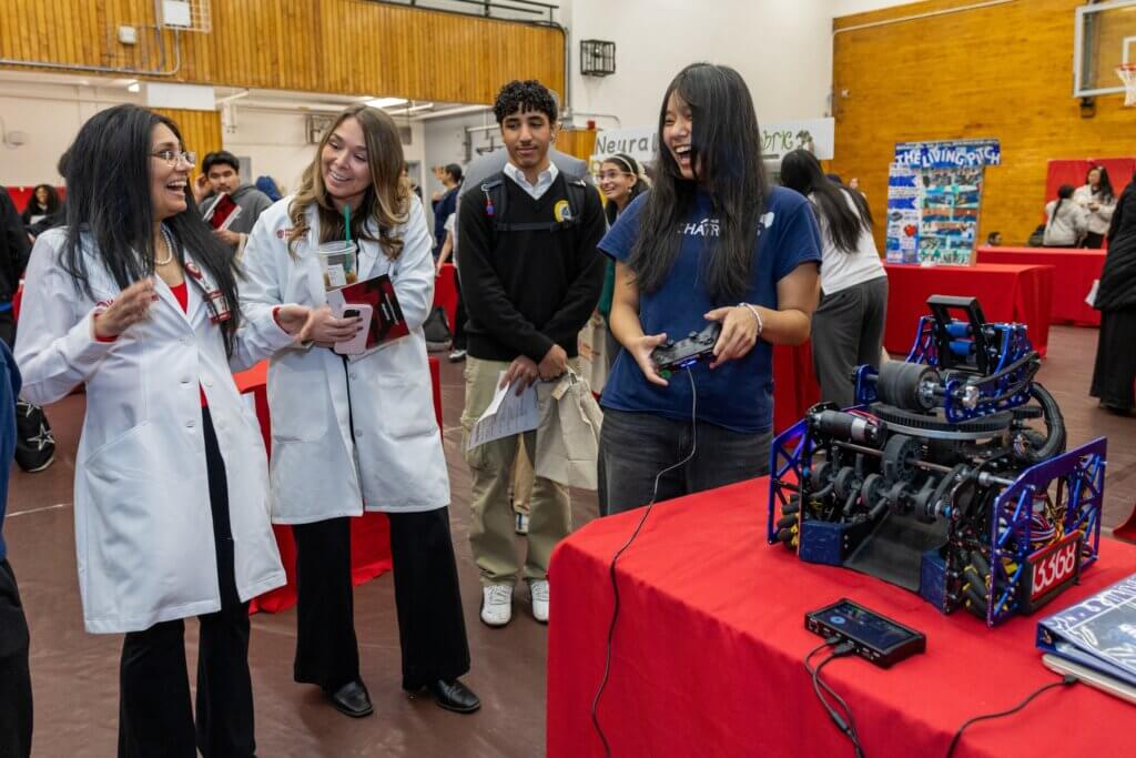 A group of students and educators interact with a robotics demonstration at a science fair in a gymnasium with red tables and posters.