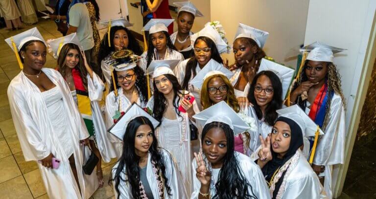 A group of female graduates in white caps and gowns pose together indoors, some smiling and making peace signs, looking up at the camera.