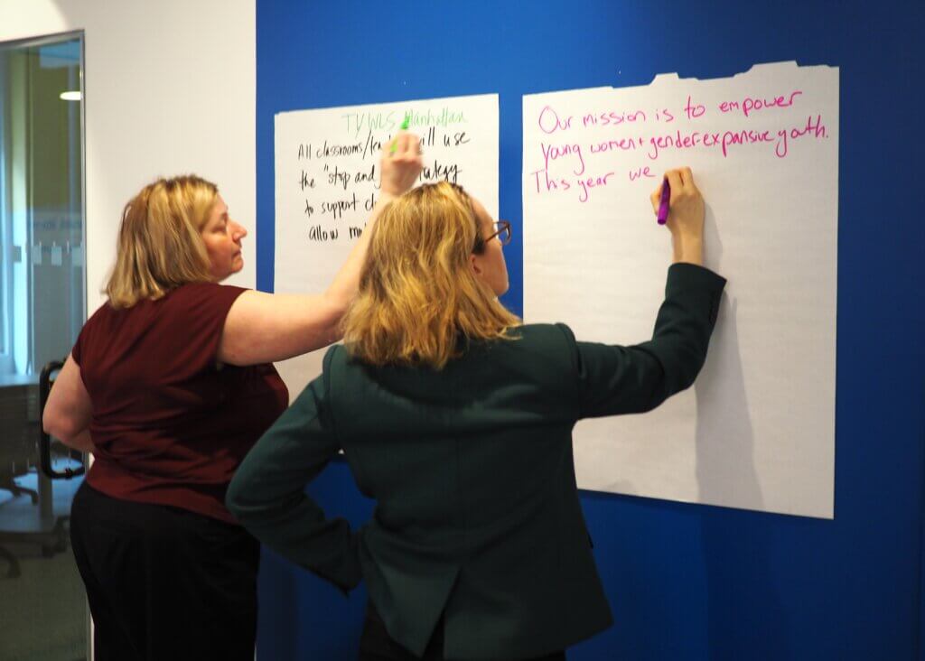 Two women write on large sheets of paper taped to a blue wall during a meeting or workshop, one using a green marker and the other a pink marker.