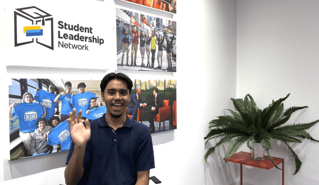 A young man sits and waves in front of a wall with Student Leadership Network signage, photo collages, and a green potted plant on a small red table.