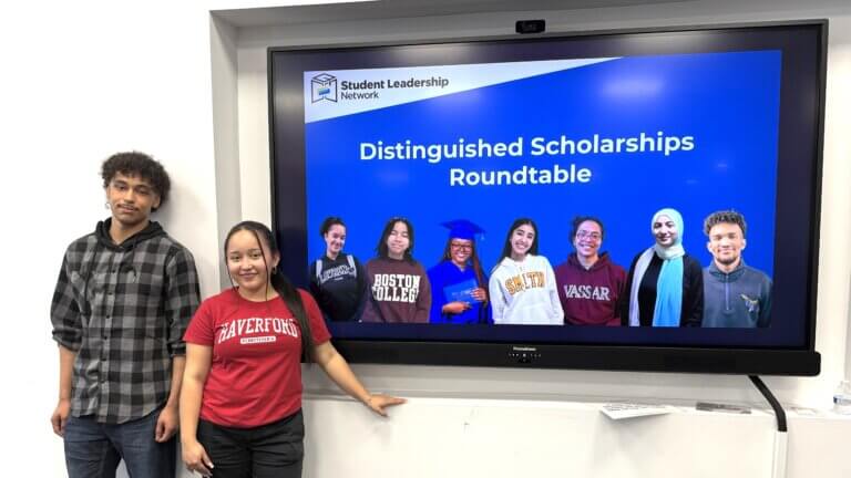 Two students stand by a screen displaying Distinguished Scholarships Roundtable, featuring images of students alongside information about college scholarships such as QuestBridge and the Posse Foundation.