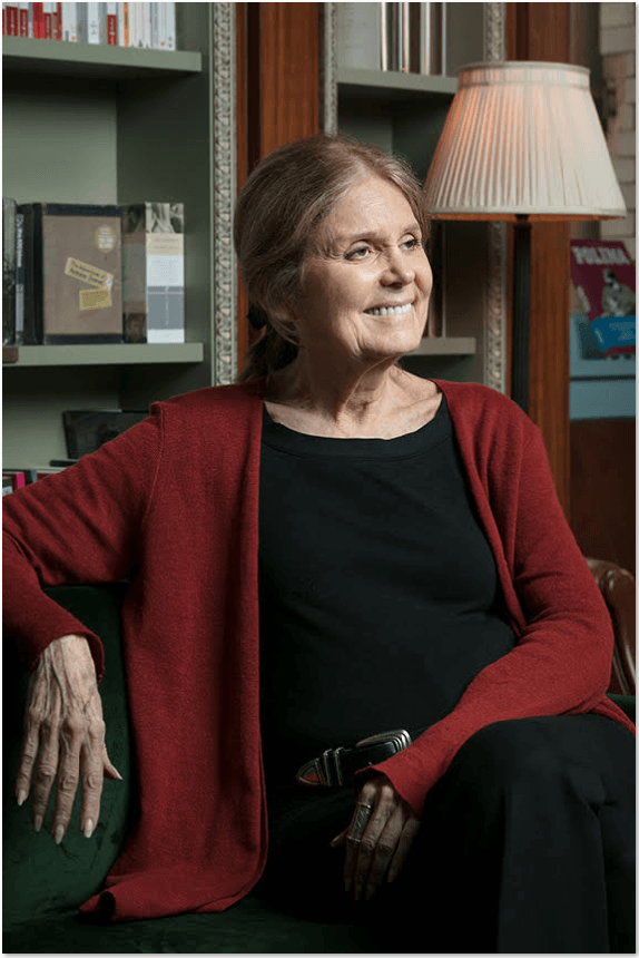 Gloria Steinem sits on a green couch, wearing a red cardigan and black clothes, smiling and looking to her left, with a bookcase in the background