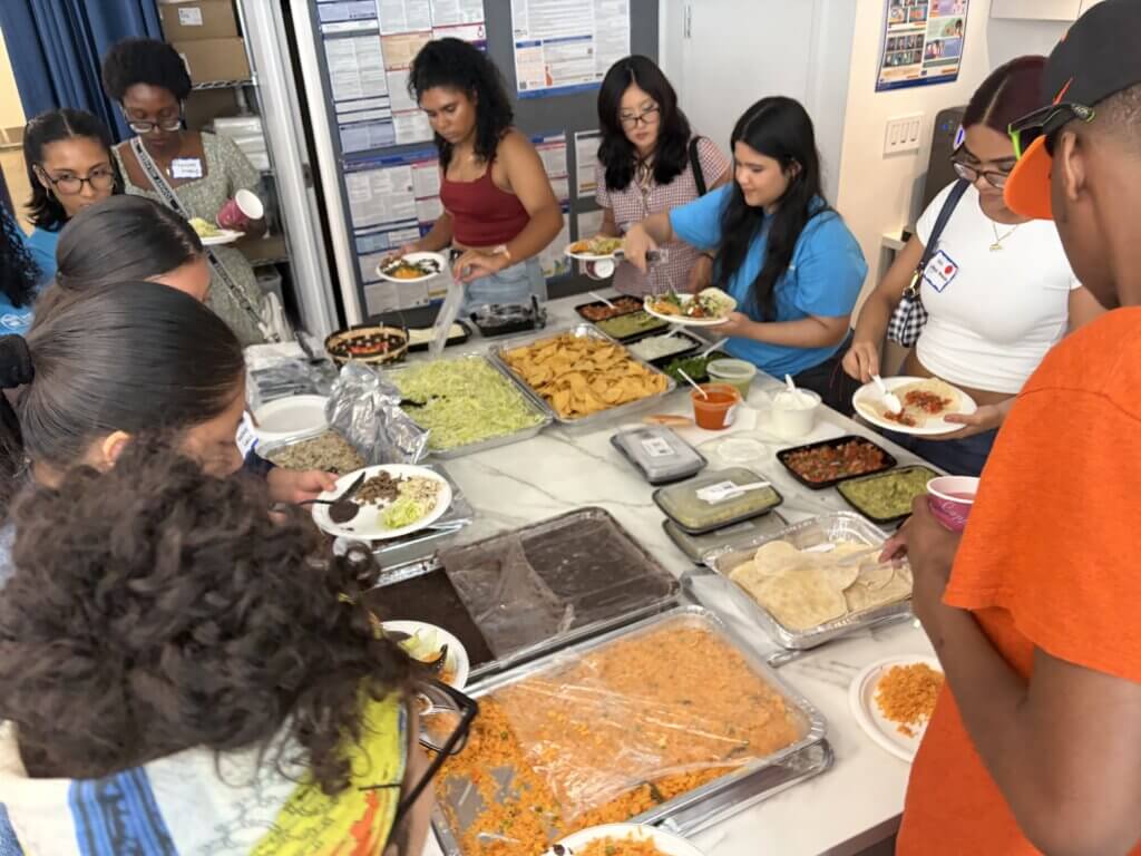 A group of people serves themselves food from a buffet-style spread of Mexican dishes in a kitchen setting.
