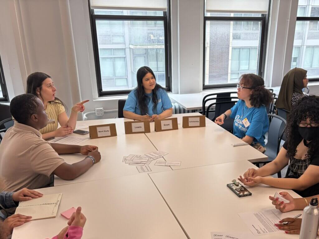 Six people sit around a table, engaged in discussion, with name tags and cards on the table in a bright room with large windows.