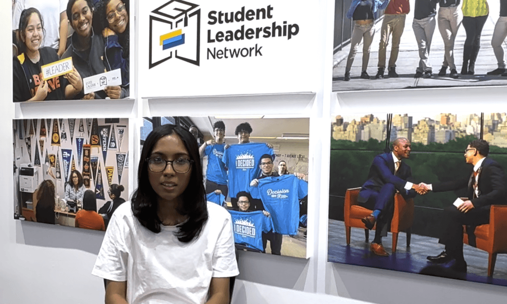 A person in a white shirt sits in front of a display featuring the Student Leadership Network logo and photos of group and professional activities.