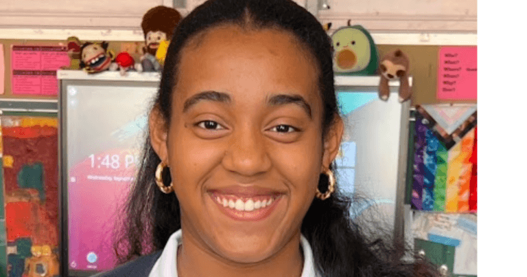 Student spotlight: Zoe Trinidad—a young person with long dark hair and gold hoop earrings—smiles in a colorful classroom filled with posters and toys, radiating a sense of community and warmth.