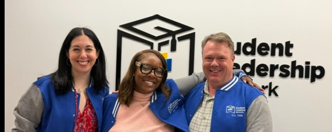 Three people in blue jackets stand smiling in front of a wall with the words Student Leadership Network and a cube logo, representing the Voices Behind the Mission.