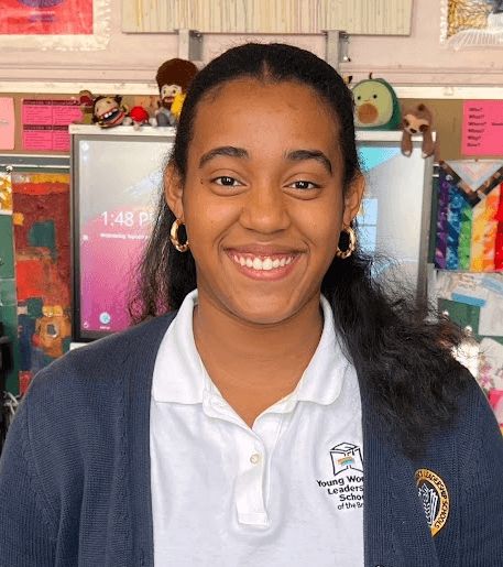 A young woman, Zoe Trinidad, in a school uniform smiles at the camera in a colorful classroom with quilts and toys in the background, embodying the spirit of community in this student spotlight.