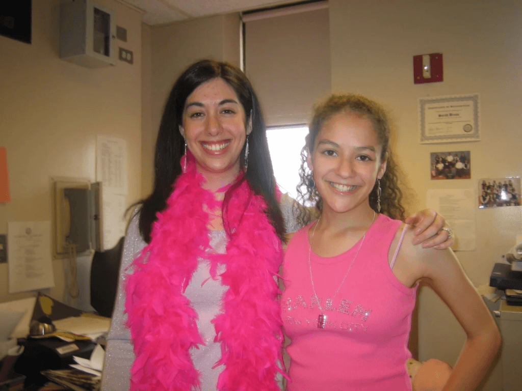 Two people stand smiling in an office, embodying the spirit of “Voices Behind the Mission.” One wears a pink feather boa and sweater, while the other sports a pink tank top. Papers and office supplies are visible in the background.