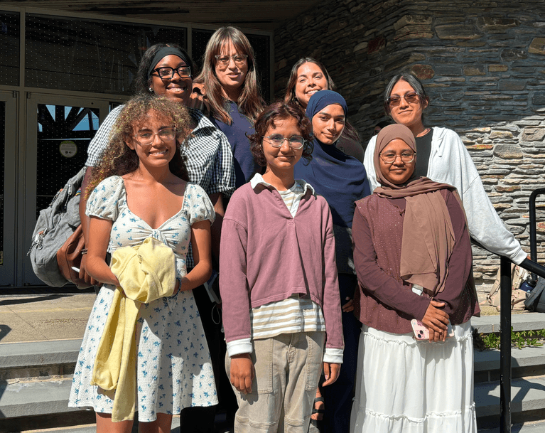 Nine people stand together on outdoor steps in bright sunlight, smiling at the camera in front of a building with stone walls and large windows, capturing a moment from their field research as part of a secondary school research program.