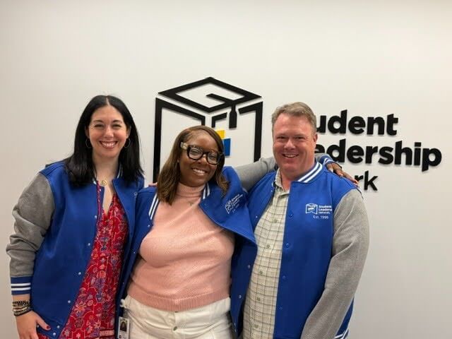 Three people wearing blue and gray jackets stand smiling in front of a Student Leadership Network sign on a white wall, representing the Voices Behind the Mission.