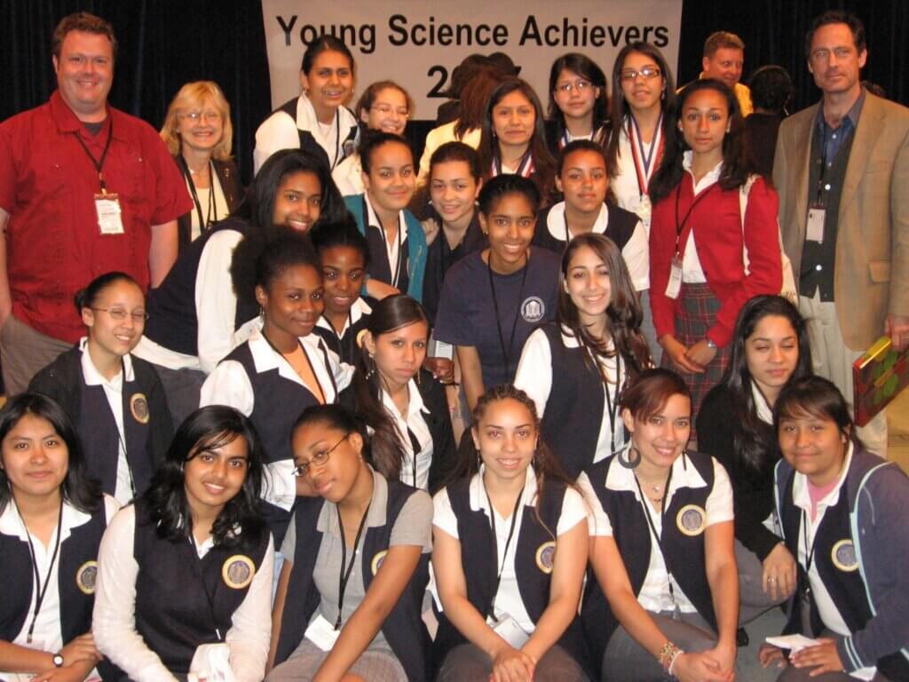 A group of young women and a few adults pose together in front of a Young Science Achievers 2007 banner, proudly representing the Voices Behind the Mission.