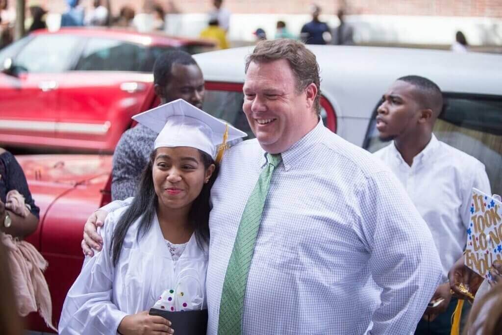 A graduate in a white cap and gown stands next to a smiling man in a dress shirt and tie outdoors, with other people and cars in the background—capturing one of the many proud moments that represent the Voices Behind the Mission.