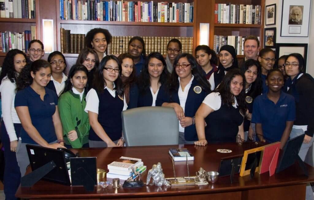 A group of students and one adult, representing the Voices Behind the Mission, pose for a photo around a large desk in a library or office with bookshelves and framed pictures on the wall.