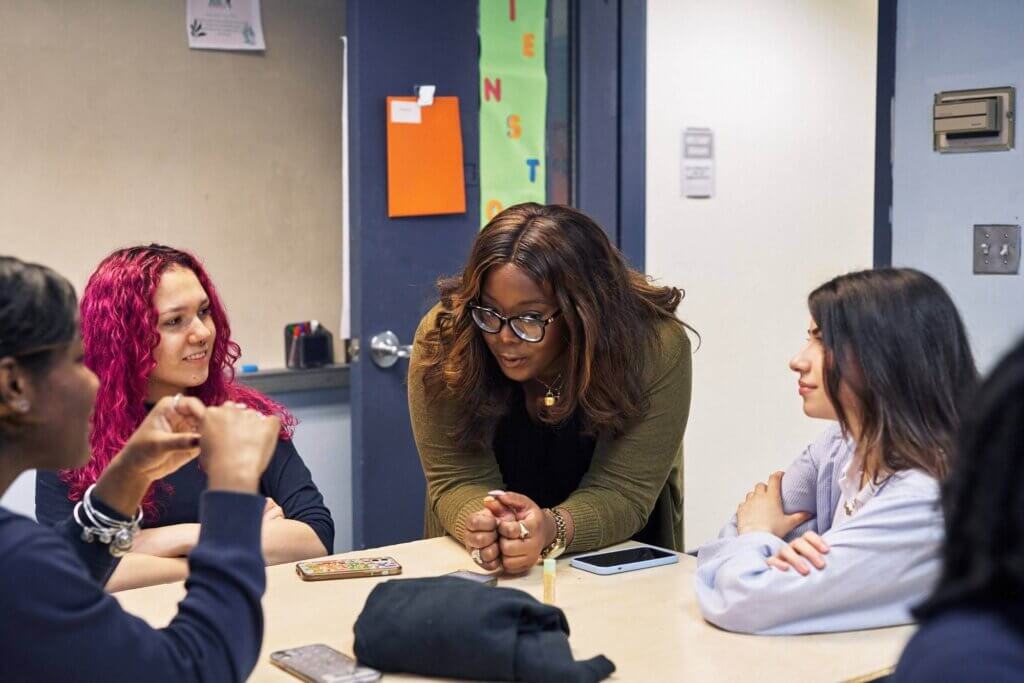 A woman stands and leans over a table, talking to a group of three seated people in a classroom setting, embodying the spirit of collaboration at the heart of Voices Behind the Mission.