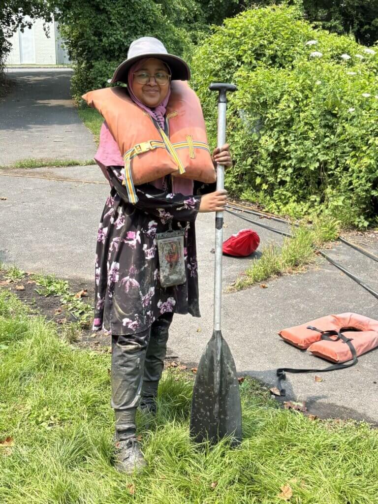 Person wearing a hat, glasses, and life jacket stands on grass holding a paddle, likely participating in a Secondary School Research activity. Another life jacket and a red bag are on the ground nearby. Green bushes and pavement are in the background.