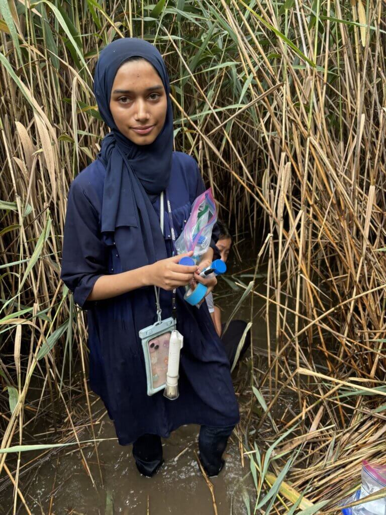 A woman wearing a dark hijab and dress stands in shallow water among tall reeds, holding plastic bags and a blue container, with a phone around her neck—possibly documenting samples for a Field Research Program or participating in Secondary School Research.