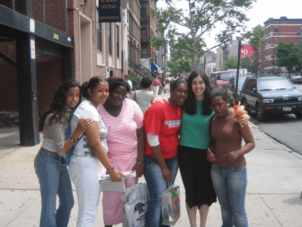 Six women, the Voices Behind the Mission, stand together on a city sidewalk, smiling at the camera. Some hold bags and gift items, while cars and buildings line the street in the background.