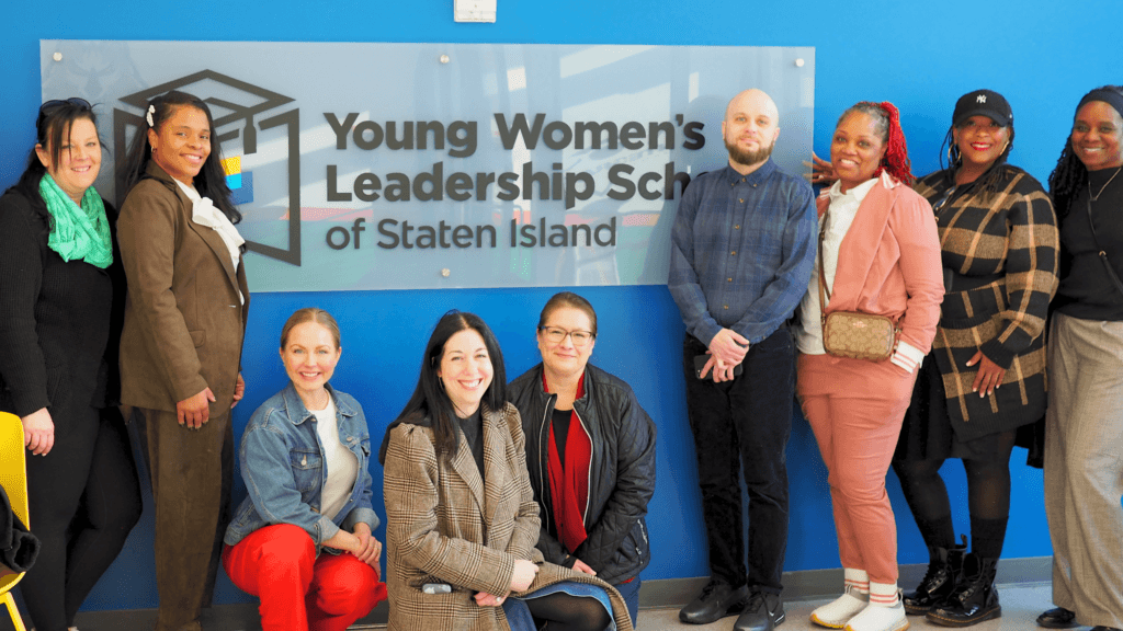 A group of ten diverse WholeGirl+ staff members pose in front of a Young Womens Leadership School of Staten Island sign on a blue wall.